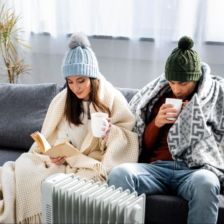 Couple sitting next to portable heater wearing beanies & blanket