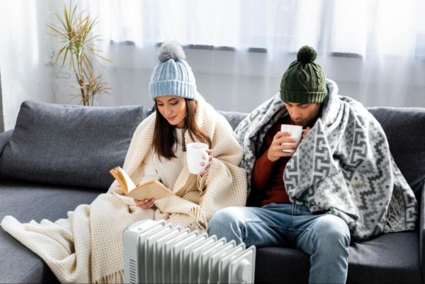 Couple sitting next to portable heater wearing beanies & blanket