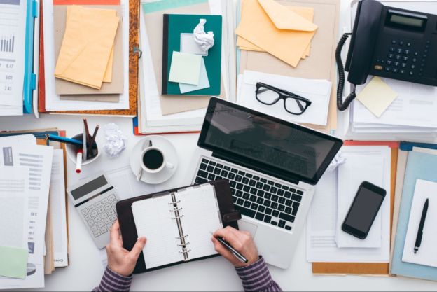 Person filling out planner next to a laptop & stack of notebooks